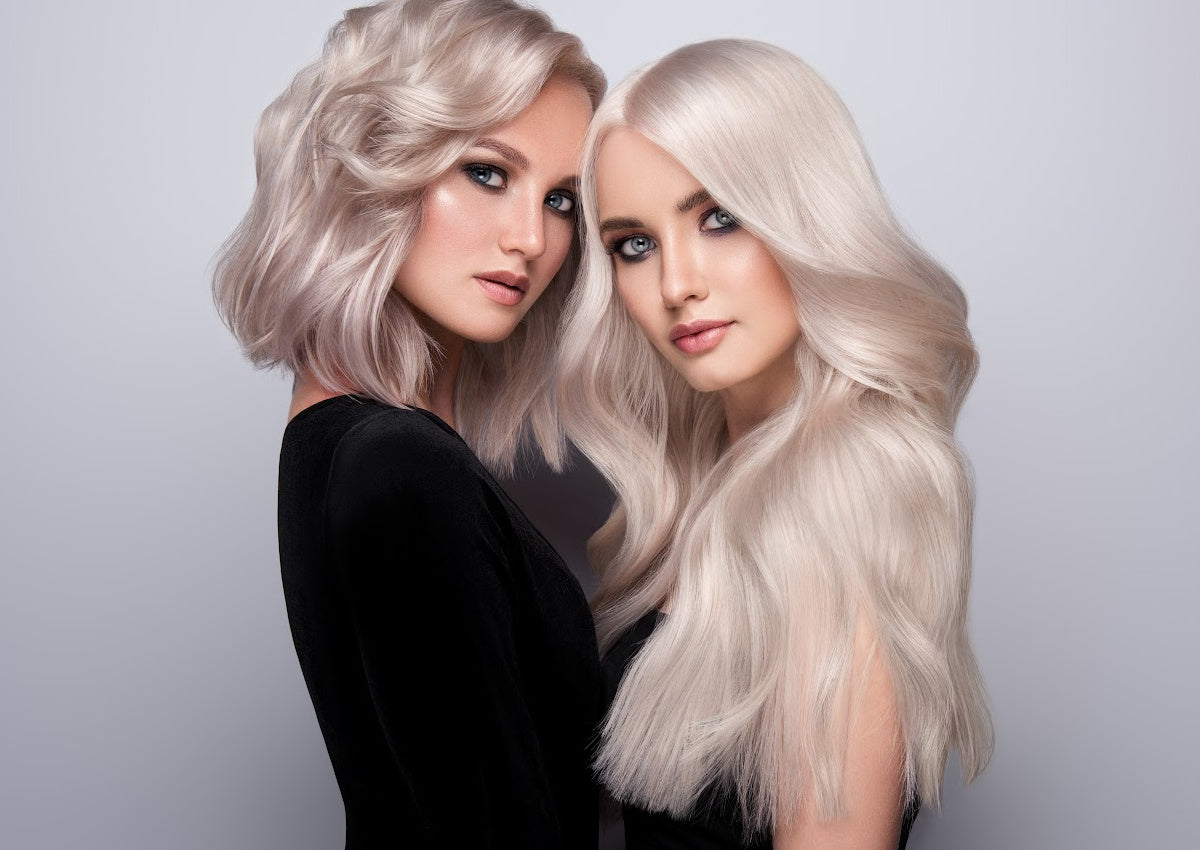Two women with platinum blonde Seamless I-Tip microlink extensions styled smooth and voluminous, part of SL Raw Virgin Hair collection.