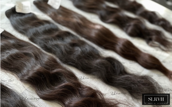 Close-up of raw human hair extensions laid out, showcasing natural black shades with slight color variations and diverse wave and curl patterns from single donors.