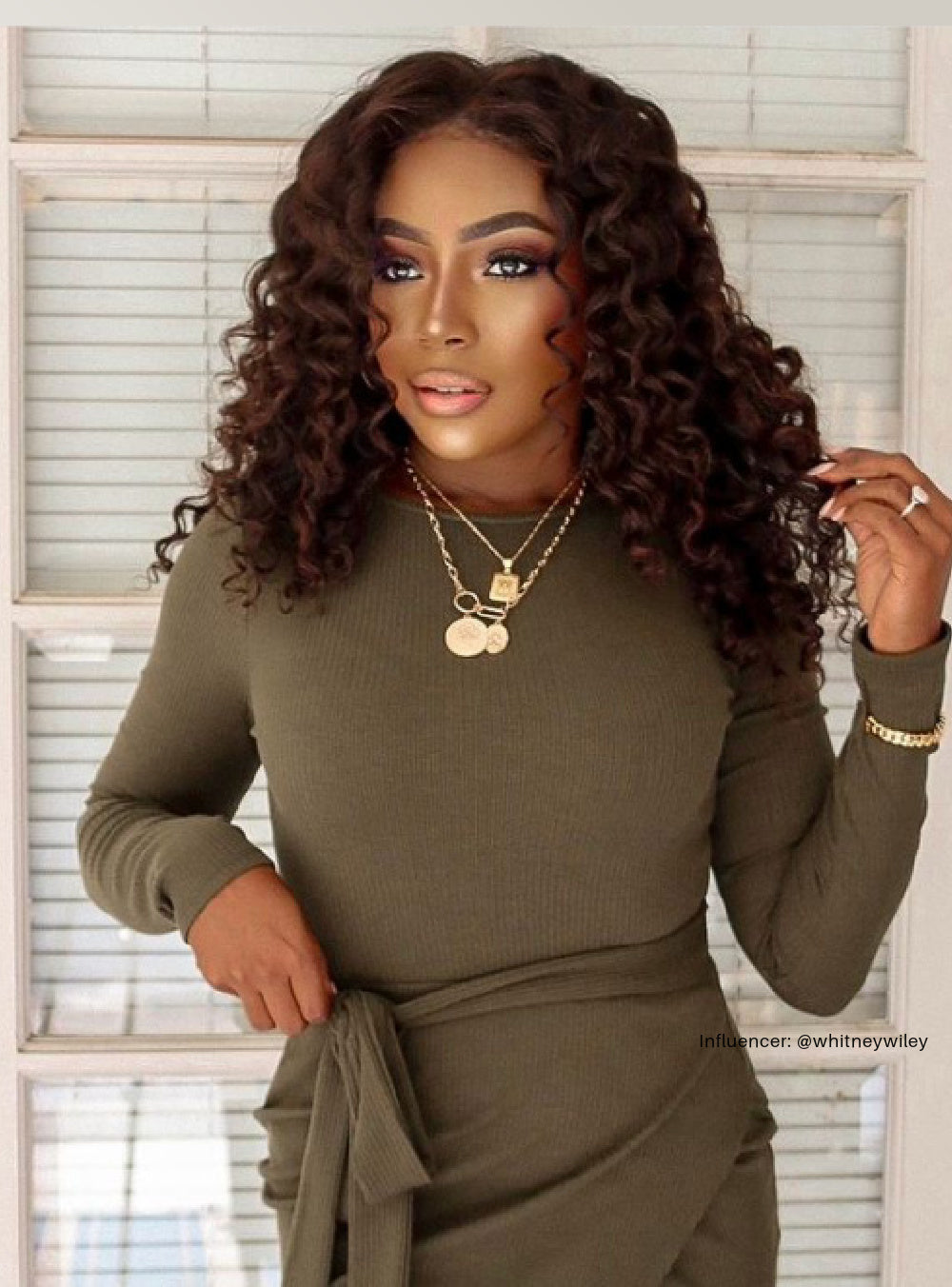 A woman wearing curly hair extensions in a green long-sleeve top with a belt, standing in front of a window.