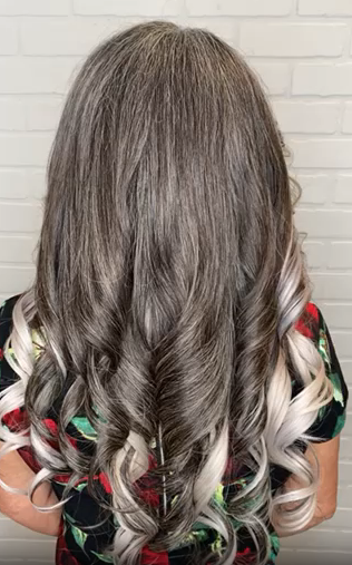 Vista trasera de una clienta con cabello humano largo gris sal y pimienta peinado en rizos sueltos mezclados con extensiones cosidas de color plateado sólido.