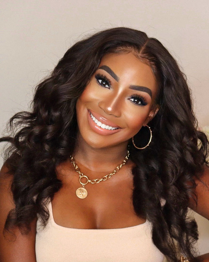 Woman with long, wavy hair wearing a gold necklace and hoop earrings against a neutral background