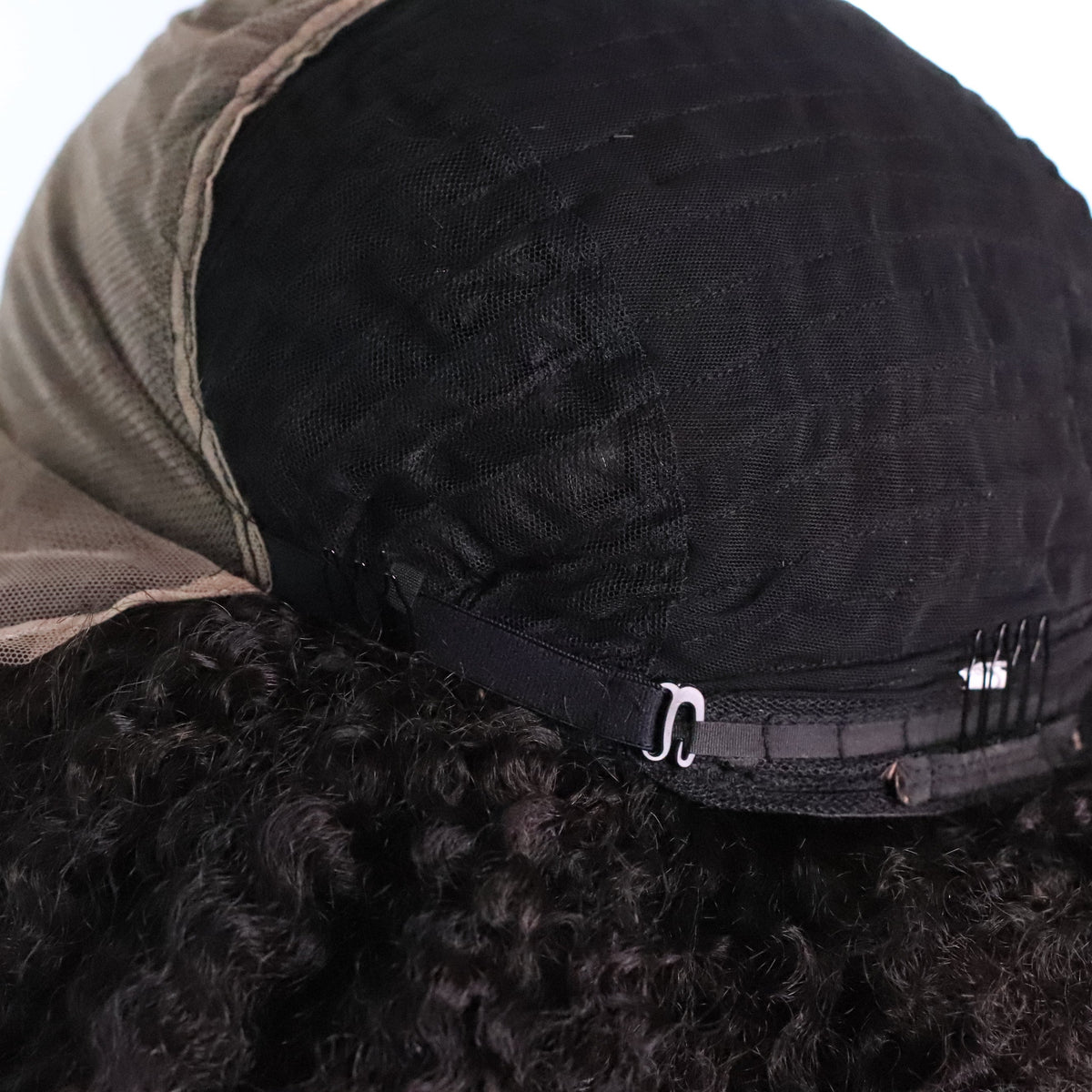 Close-up back view of a black wig with a beige lace hairline and a black elastic band with metal clips, shown on a white background.